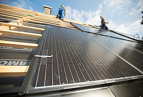 Installation von Photovoltaik auf einem Dach: Module auf Dachlatten von unten fotografiert, oben Schornstein und zwei Handwerker
